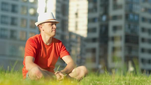 Un joven con un sombrero descansando sobre la hierba en un parque de la ciudad, la cámara rastreando — Vídeos de Stock