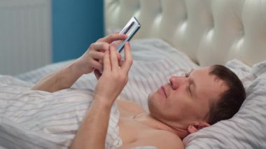 Sabahleyin, genç bir adam yatağında yatarken haberlere cep telefonundan bakıyor.