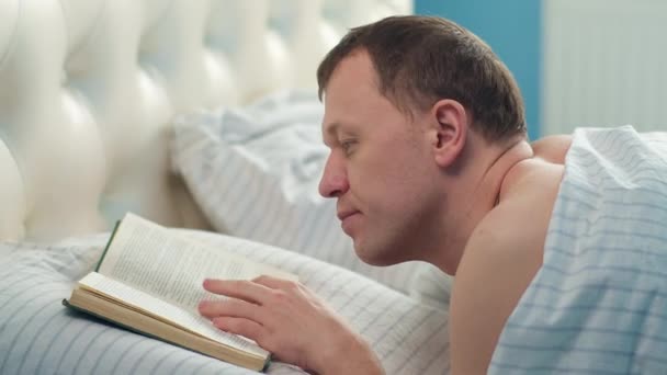 Homem lendo livro enquanto deitado na cama pela manhã, movimento da câmera, conceito de relaxamento — Vídeo de Stock