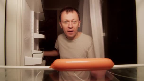 Hombre hambriento toma una salchicha de la nevera estante, toma un bocado de ella — Vídeos de Stock