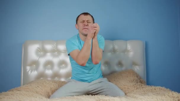 Attractive young man having fun sitting on the bed, raised his arms and dancing while sitting in front of the camera — Stock Video