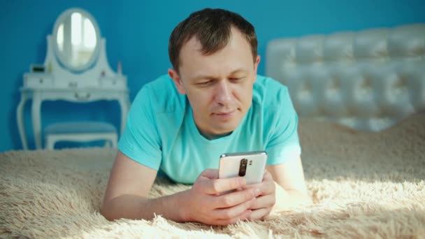 Молодой человек лежит в спальне на кровати с мобильным телефоном в руках — стоковое видео