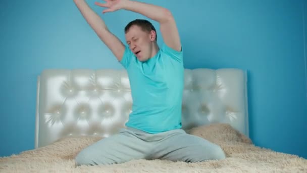 Young man having fun while sitting on the bed, raised his hands and dances, sitting of the camera — Stock Video
