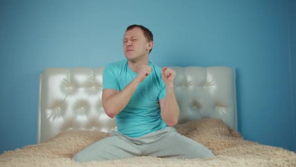 Man having fun while sitting on the bed, raised his hands and dancing, sitting in front of camera — Stock Video