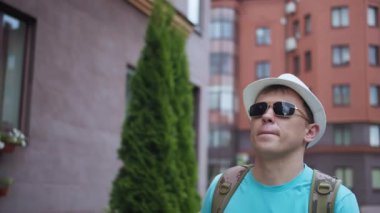 Erkek turist şehirde şapka ve güneş gözlüğü takarak dolaşıyor, kamera takibi yapıyor.