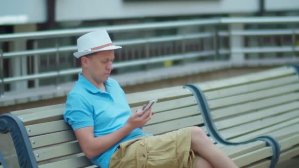 Jovem sentado em um banco da cidade com um telefone celular em suas mãos, rastreamento de câmera — Vídeo de Stock