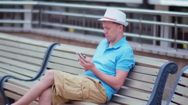 Young man in a hat sits on a city bench with a mobile phone in his hands, camera tracking — Stock Video