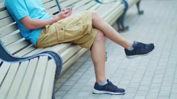 Włochate nogi mężczyzny w szortach siedzącego na ławce z telefonem komórkowym, śledzącego kamery — Wideo stockowe
