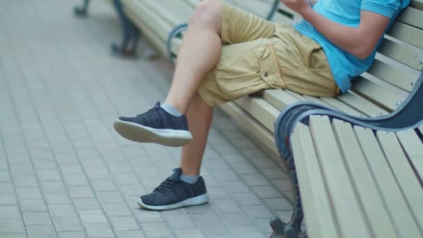 Legs of man in shorts sitting on city bench with mobile phone tracking camera — Stock Video