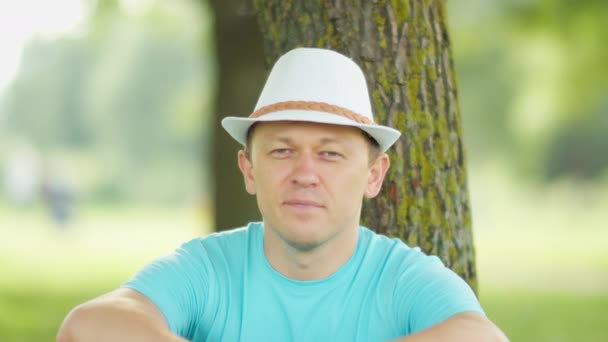 Retrato de um jovem com um chapéu branco, sentado debaixo de uma árvore e olhando para a câmera, close-up — Vídeo de Stock