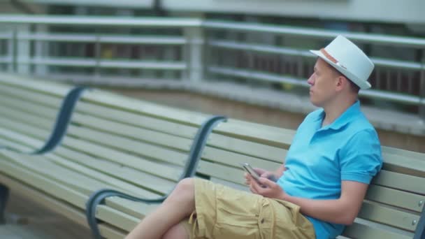 A man in a white hat is resting while sitting on a city bench, tracking a camera — Stock Video