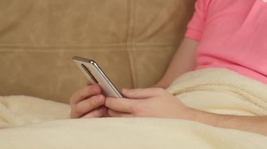 Pembe tişörtlü bir adam kanepenin üzerinde, battaniyenin altında cep telefonuyla oturuyor.