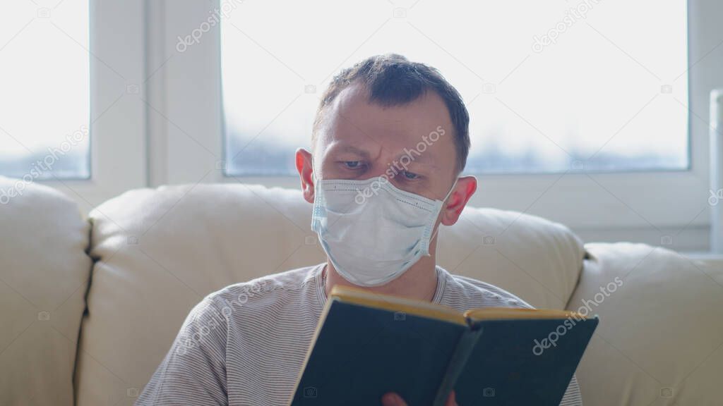 Attractive man in mask with a book in his hands looks at the camera while sitting on a sofa