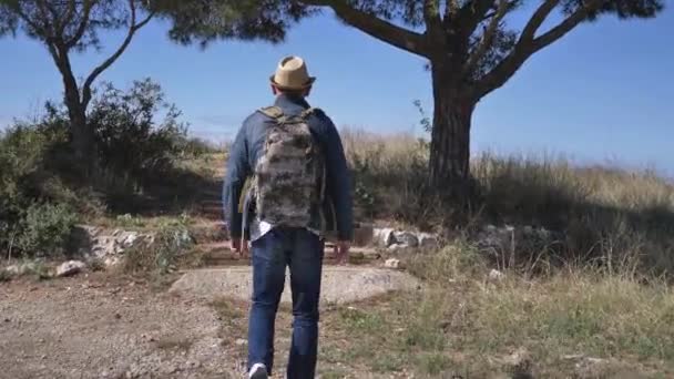 Un hombre viajero en un sombrero y con una mochila camina por el sendero y lo llama — Vídeos de Stock