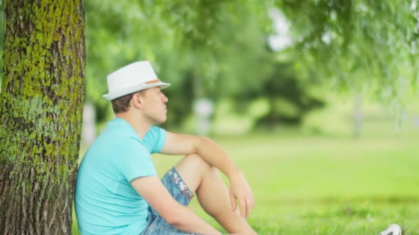 Vista lateral de un hombre cansado con un sombrero blanco descansando a la sombra de un árbol en el parque — Vídeos de Stock