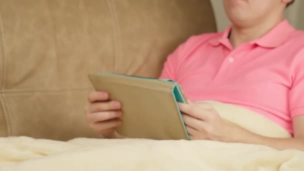 Rózsaszín pólós férfi ül a kanapén a takaró alatt egy tablettával. — Stock videók