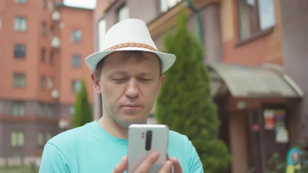 A young man in a hat stands on the street of the city, thoughtfully examines the information in his mobile phone — Stock Video