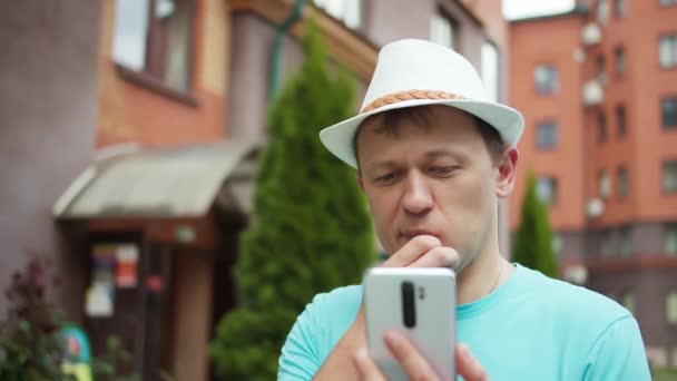 Young man in a hat stands on the street of the city, thoughtfully examines the information in his mobile phone, tracking camera — Stock Video