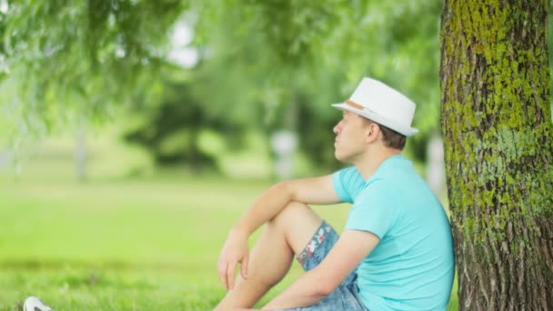 Vista laterale di un uomo stanco con un cappello che riposa all'ombra di un albero nel parco — Video Stock