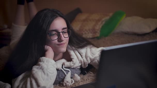 Menina com óculos à noite lê a notícia em um laptop está na cama — Vídeo de Stock