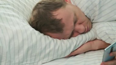 Close-up of a man in bed with a mobile phone, head on the pillow visible bald patches, camera tracking