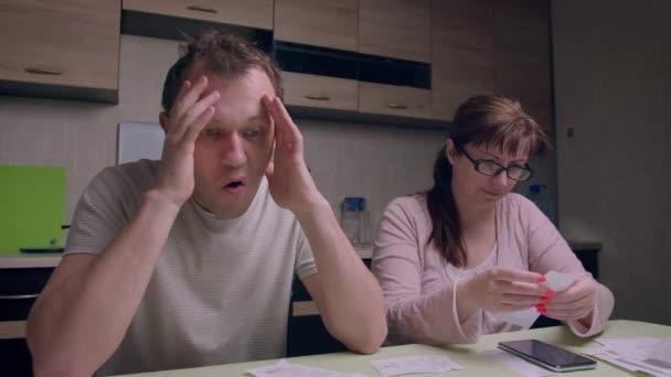 Сім'я сидить за кухонним столом увечері і перевіряє квитанції про покупку, чоловік перебуває в емоційному стресі — стокове відео