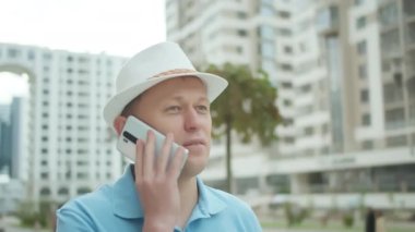 Beyaz şapkalı genç adamın portresi cep telefonuyla konuşuyor, şehir geçmişi, kamera takibi