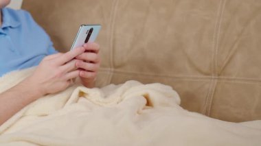 Elinde cep telefonu olan bir adam kanepeye oturur. Battaniyeyle kaplıdır.