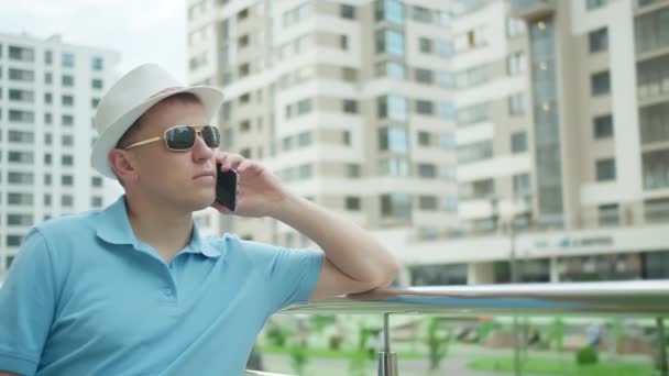 Joven hombre confiado en sombrero hablando en el teléfono móvil, mientras que de pie cerca de barandilla valla — Vídeos de Stock