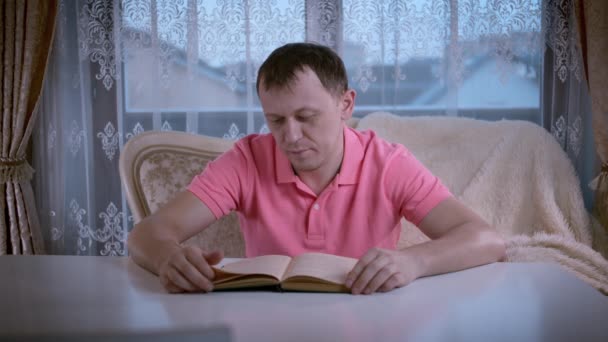 Mannen i rosa t-shirt läser en bok medan han sitter på soffan på kvällen — Stockvideo
