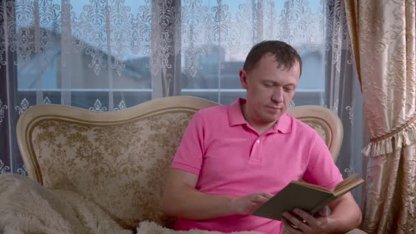 Férfi olvasás egy könyvet, miközben ül este a kanapén, ablak háttér — Stock videók
