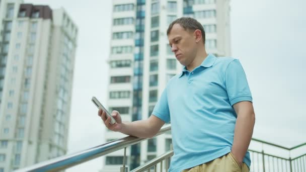 Retrato de um jovem com um telefone celular em suas mãos, fica nas escadas, edifícios de fundo — Vídeo de Stock