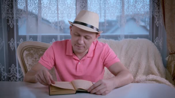 Un hombre con sombrero por la noche lee un interesante libro sentado en una mesa — Vídeo de stock