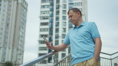 Elinde cep telefonu olan bir adamın portresi, merdivenlerde duruyor, binalar arka planda.