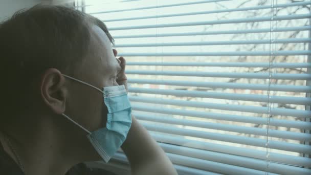 Man in medical mask looks out the window through the blinds, self-isolation — Stok video