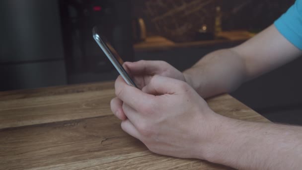Freelancer at home sits at a wooden table, reads a phone message or downloads an application for his mobile phone — Stock Video
