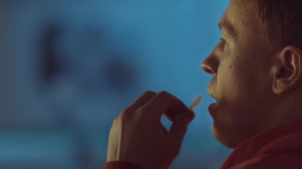 Retrato de cerca de un hombre que come patatas fritas y mira la televisión a la luz de la noche, reflejo de color — Vídeos de Stock