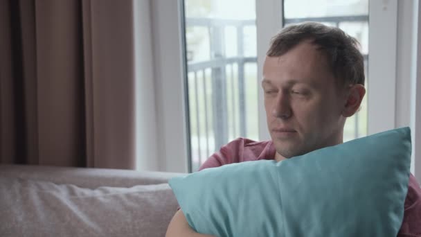 Portrait of a young man immersed in his thoughts sits on a sofa clutching a pillow to himself — Stock Video