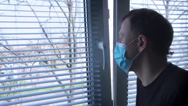 Young man in medical mask stands by the window with blinds and sadly looks out into the street — Stock Video