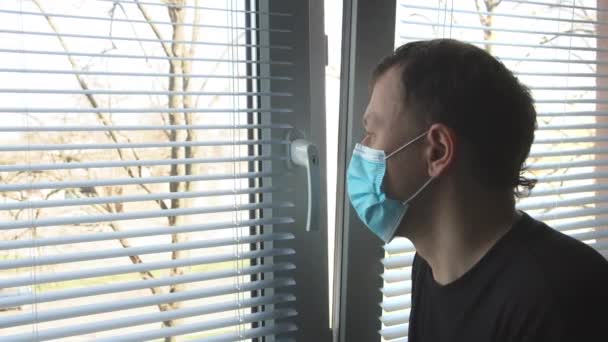 Em um dia ensolarado, um homem fica em uma janela com persianas e olha tristemente para a rua — Vídeo de Stock