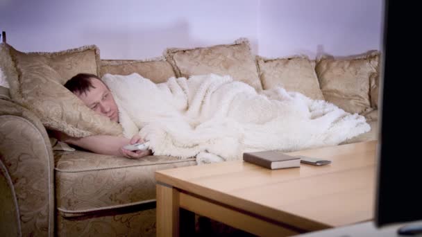 Um homem está coberto com um cobertor em um sofá, muda de canais de TV, iluminação noturna — Vídeo de Stock