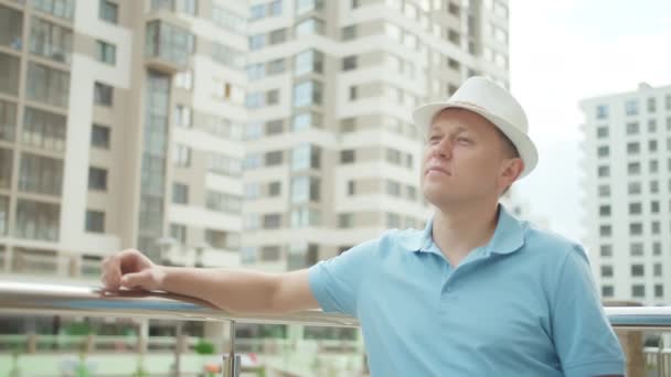 Tourist in a hat stands at a metal fence, put his hand on the railing, looks around — Stock Video