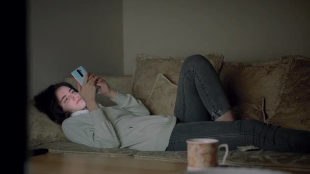 Ein einsames Mädchen verbringt den Abend allein mit dem Handy auf der Couch liegend — Stockvideo