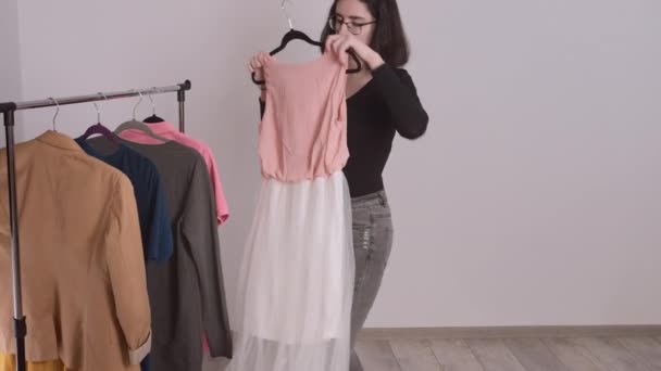 Hermosa joven de pie en el perchero de ropa, recogiendo la ropa, armario básico. — Vídeos de Stock