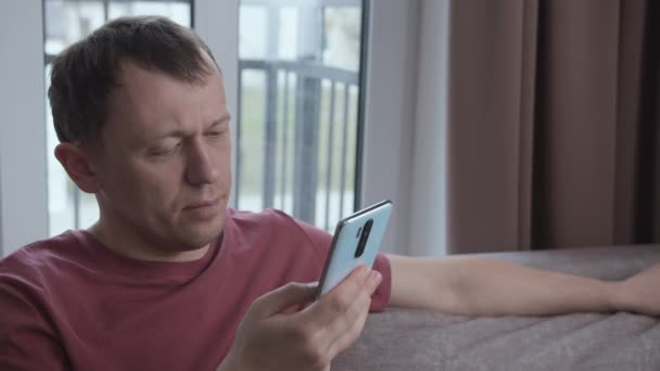 Homem lendo mensagens de texto, navegando na Internet no smartphone, sentado no sofá — Vídeo de Stock