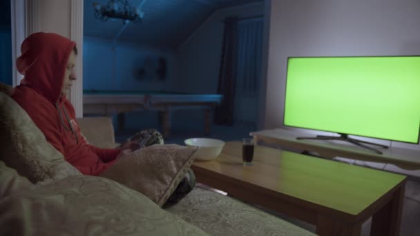 Mann sitzt mit Handy auf Sofa vor grünem Fernseher — Stockvideo