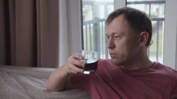 Man med ett seriöst uttryck dricker från ett glas som sitter på sofffönstret bakgrund — Stockvideo