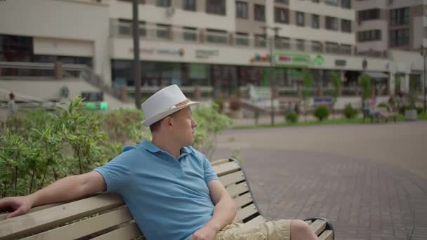 Человек в шляпе сидит на городской скамейке, отдыхает во время прогулки по городу, движения камеры — стоковое видео