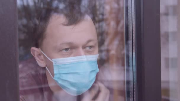 Uomo con una maschera medica dietro il vetro, guarda fuori dalla finestra sulla strada, resta a casa, localizza le telecamere — Video Stock