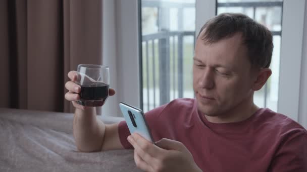 Portret van een serieuze man die op de bank zit met een glas drinken en boodschappen lezen in een mobiele telefoon — Stockvideo